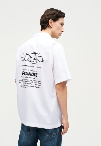 Άνδρας που φοράει λευκό πουκάμισο με κοντό μανίκι, με μαύρο γραφικό του Snoopy και το κείμενο Peanuts στην πλάτη, σε συνδυασμό με μπλε τζιν, στραμμένος στο πλάι.