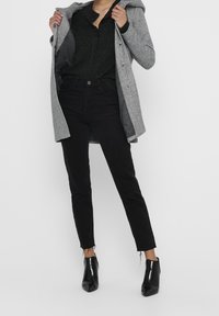 Grauer Mantel mit Kapuze, schwarzer, geknöpfter Pullover und schwarze Jeans mit fransigen Säumen, kombiniert mit eleganten schwarzen Stiefeletten.