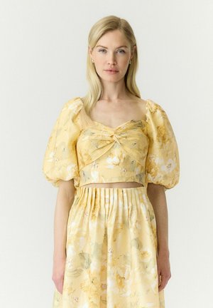 Femme blonde portant un crop top jaune à manches bouffantes fleuri et une jupe taille haute assortie, debout devant un fond clair uni.
