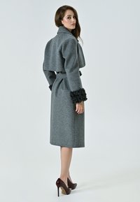 Cappotto grigio in lana con un design corto, ampio colletto e polsini in pelliccia nera testurizzata. Il cappotto presenta una cintura e arriva sotto il ginocchio.