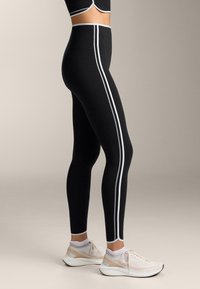High-waisted black leggings with white side stripes, made of stretchy fabric. Paired with light athletic shoes featuring pastel accents.