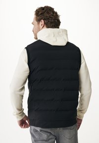 Muž s kudrnatými vlasy, oblečený v černé prošívané vestě přes krémovou mikinu a šedé džíny, viděný zezadu na bílém pozadí.