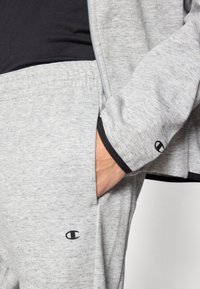 Sudadera y pantalones deportivos grises hechos de tela suave. Presentan un cierre delantero con cremallera, detalles negros en las mangas y un logo en los pantalones deportivos.