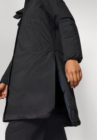 Zwarte geïsoleerde parka met een gladde textuur, voorzien van drukknoopsluitingen, zijslitsen en een ritssluiting aan de binnenkant. Een arm is zichtbaar die de jas vasthoudt.