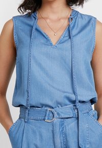 Denim ärmelloses Top mit V-Ausschnitt, Frontnähdetail und Kordelzugakzenten in einem hellblauen Farbton. Glatte Stoffstruktur.
