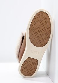 Semelle d'une botte UGG marron avec une semelle extérieure en caoutchouc beige et un motif de semelle géométrique, montrant un doublage en fourrure douce à l'intérieur de la botte.