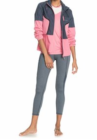 Giacca antipioggia leggera in blu navy e rosa, abbinata a una maglietta rosa e leggings grigi con cuciture a contrasto. Posizione a piedi nudi.