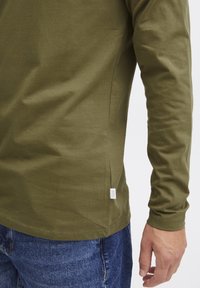 Olijfgroene katoenen shirt met lange mouwen en een ronde zoom, voorzien van een label opzij, gecombineerd met blauwe spijkerbroek. Soepele textuur en getailleerd ontwerp.