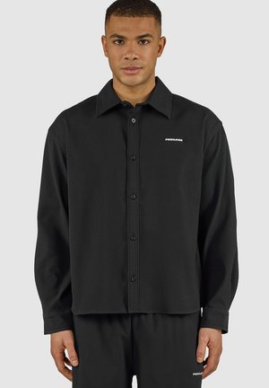 Man wearing black ribbed long-sleeve button-up shirt and matching pants with small white "PEGADOR" logo on chest and thigh.