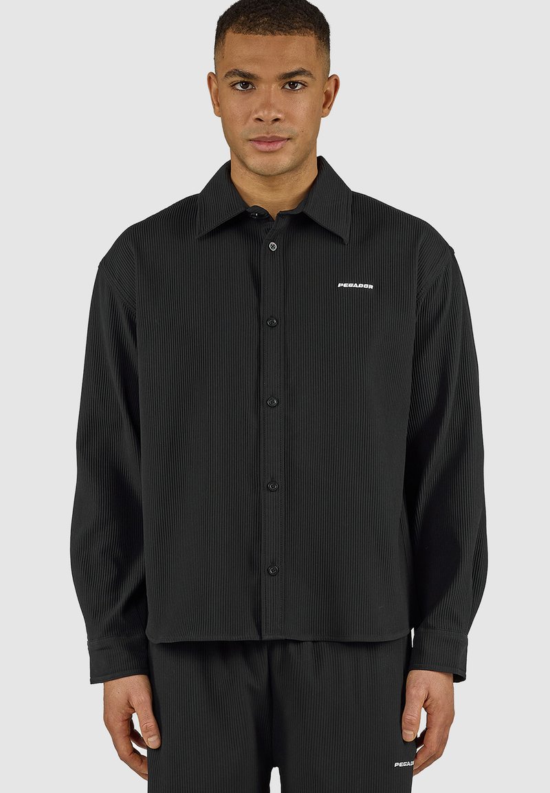 Hombre con camisa negra a rayas acanaladas de manga larga abotonada y pantalones a juego con pequeño logo blanco "PEGADOR" en el pecho y el muslo.