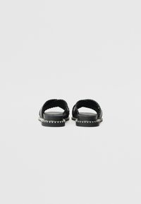 Black sandals with a flat sole, featuring wide woven straps at the upper and white stitching along the sole edge.