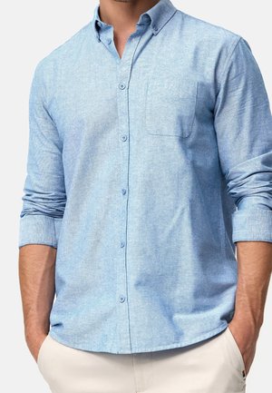 Man draagt een lichtblauw overhemd met lange mouwen en opgerolde manchetten en een beige broek, handen in de zakken.