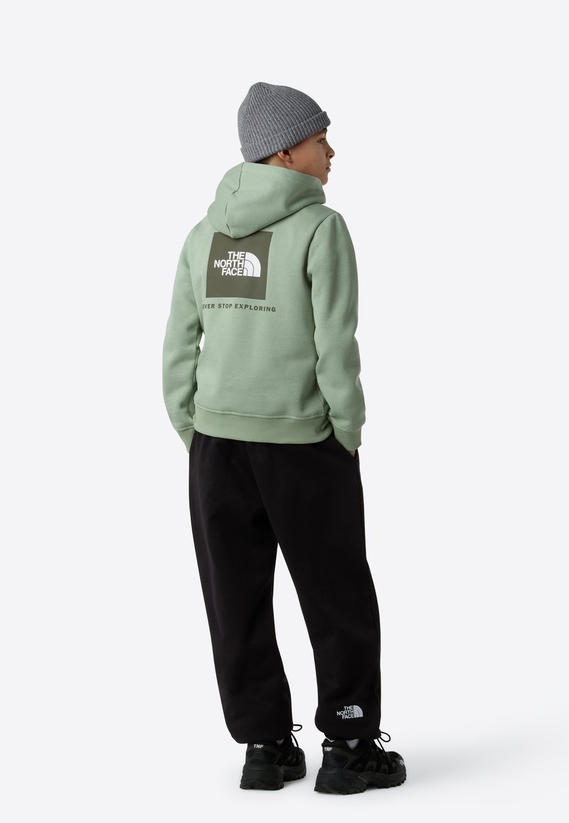 Grønn hettegenser med en stor logo på baksiden, kombinert med svarte joggebukser og svarte sko, med en teksturert grå lue.