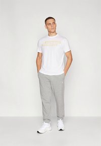 Bílé tričko s krátkým rukávem s žlutým grafickým potiskem, doplněné o šedé tepláky a bílé tenisky. Jednoduché a neformální sportovní oblečení.