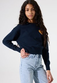 Pull marine à col rond, avec des poignets côtelés et un petit motif de cœur jaune sur la poitrine, associé à un jean taille haute bleu clair.