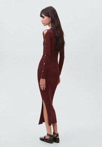 Rochie tricotată burgundy cu dungi, cu mâneci lungi, decupaje pe umeri, detalii cu nasturi și fante laterale. Asortată cu sandale negre cu curea.