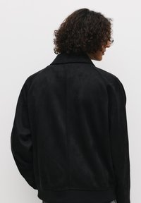 Giacca in suede nera con una texture morbida, dotata di colletto e orlo a coste. La parte posteriore presenta un design semplice senza ulteriori dettagli.