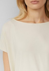Vrouw met lichtblond haar, gekleed in een eenvoudig off-white top met korte mouwen en een brede hals.