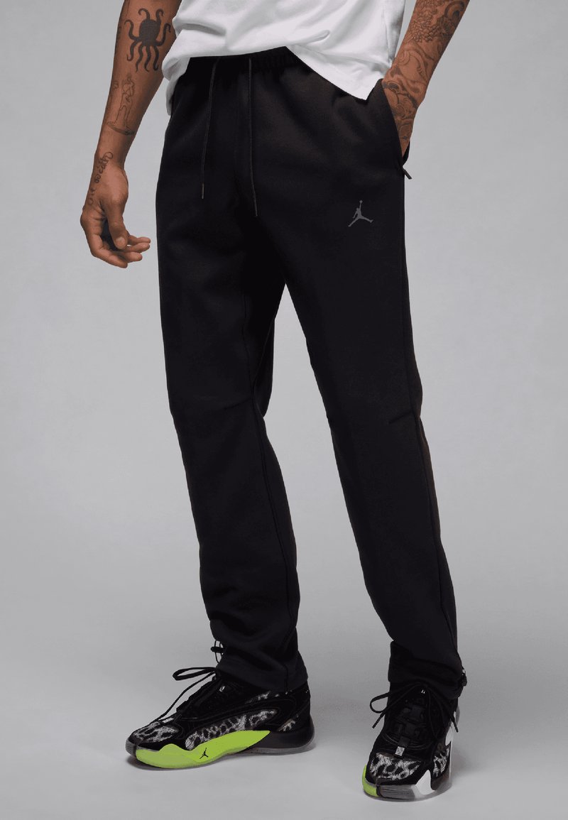 Zwarte sportieve broek met een relaxte fit, voorzien van een subtiel logo en een elastische tailleband, gecombineerd met zwarte sneakers met neongroene accenten.