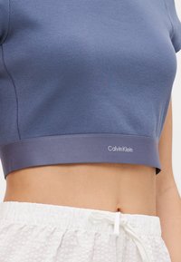 Modrý žebrovaný crop top s kontrastním pasem, na kterém je logo "Calvin Klein". Doplněno o bílé šortky s texturou.