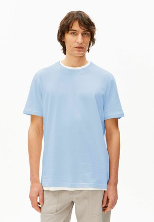 Junger Mann mit mittellangen braunen Haaren, der ein hellblaues Kurzarmhemd über einem weißen Unterhemd und beige Hosen trägt, vor weißem Hintergrund stehend.