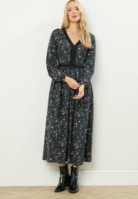 Vestido de manga larga con escote en V, cuerpo negro y estampado floral en tonos de rosa y verde sobre un fondo oscuro. La modelo lleva botas negras hasta el tobillo.