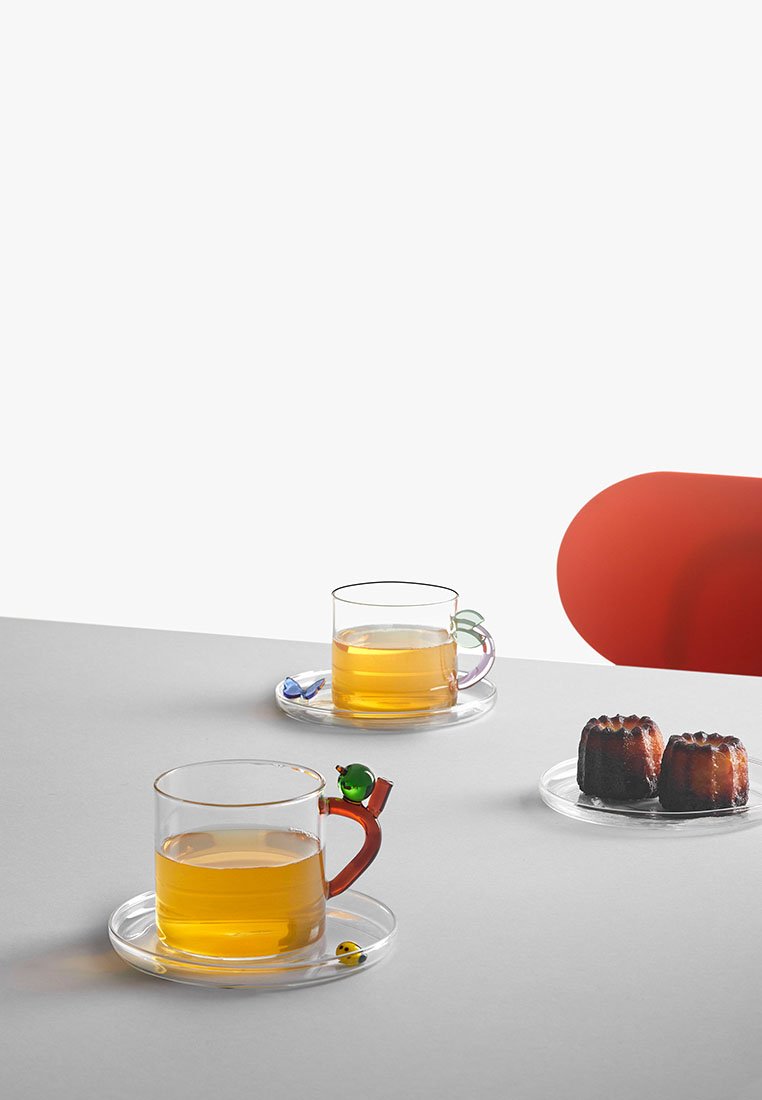Dwa przezroczyste szklanki z bursztynową herbatą na spodkach, każda z kolorowymi uchwytami w kształcie ptaków, a także talerz z dwoma ciemno-brązowymi wypiekami na szarym stole.