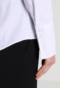 Weißes Langarmhemd mit glatter Textur, das einen abgerundeten Saum und zwei Knopfmanschetten aufweist, kombiniert mit schwarzem Stoff darunter.