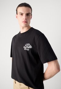 Black cotton t-shirt, relaxed fit, featuring white graphic print of a car and text "Hugo Garage" on the upper left chest.