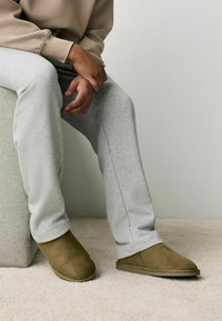 Brune ruskinds slippers med en glat overflade, iført lysegrå sweatpants, siddende på et beige tæppe ved siden af en struktureret grå overflade.