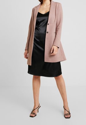 Femme portant une robe nuisette en satin noir, un blazer rose clair et des sandales à talons noires à lanières, debout devant un fond clair uni.
