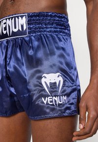 Κοντινό πλάνο ενός ατόμου που φοράει σκούρο μπλε σατέν σορτς πυγμαχίας με λευκό λογότυπο Venum στο μηρό και στη ζώνη μέσης.