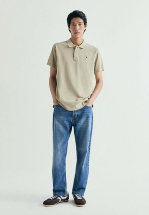 Joven de pie con las manos en los bolsillos, llevando una camisa polo beige, vaqueros azules y zapatillas negras sobre un fondo claro y sencillo.