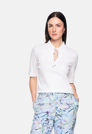 Woman with straight dark hair wears a white short-sleeve top with front tie and light blue floral patterned pants, hands in pockets.