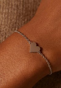 Zilveren kettingarmband met een gepolijste hartbedel, tegen een gestructureerde, tankleurige huidachtergrond.