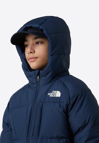 Tumesinine isoleeritud sulejope kõrge kraega, lukuga ja The North Face logo rinnal. Omadusteks on kapuuts ja polsterdatud tekstuur.