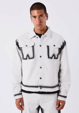 Chaqueta de mezclilla blanca con acentos que parecen estar pintados en negro, con cierres de botón y dos bolsillos en el pecho. Abertura y puños desgastados.