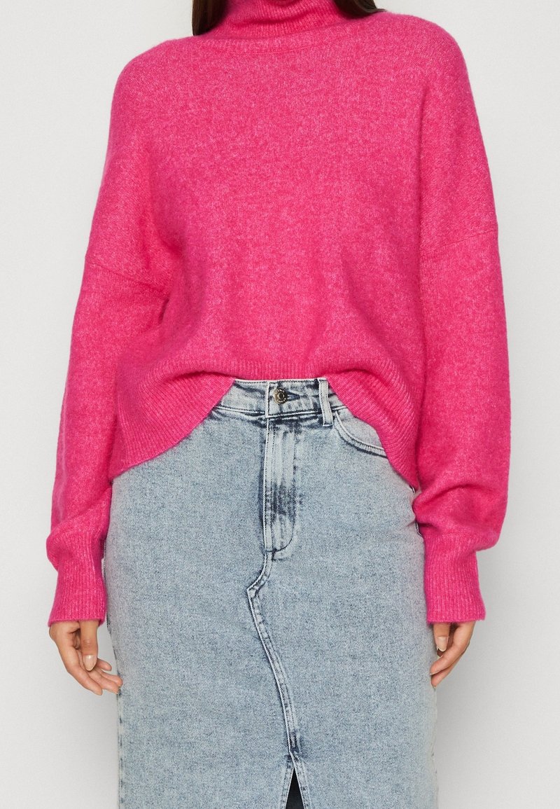 Svetle růžový oversized rolák vyrobený z měkkého materiálu, v kombinaci se světle modrou džínovou sukní s předním rozparkem a bledými detaily.