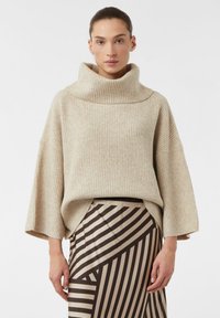 Beżowy sweter prążkowany z szerokim dekoltem w łódkę i luźnymi rękawami, zestawiony ze spódnicą w wzory z ukośnymi brązowymi i kremowymi paskami.