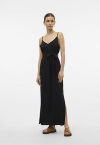 Svart maxiklänning med tunna remmar, rynkad bodice och dragsnodd i midjan. Har en sidoklyfta och flödig tyg. Buren med bruna sandaler.