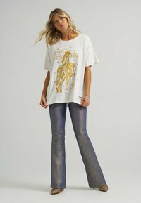 Hvid oversized t-shirt med guld grafisk print af en cowboy på en hest, parret med metallisk blå flared jeans og glitrende højhælede sko.