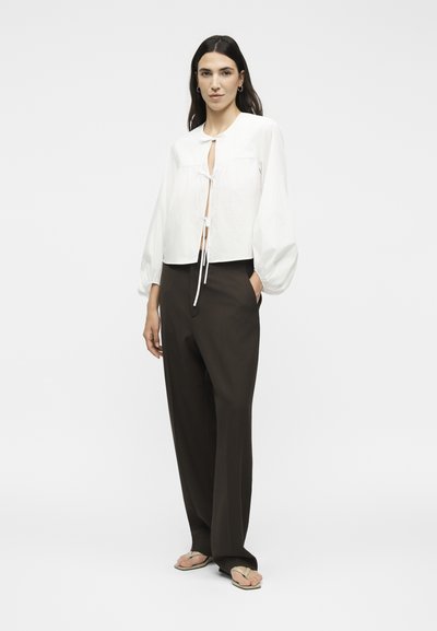 Γυναίκα που φοράει λευκό μπλουζάκι με μακριά μανίκια και κορδόνια μπροστά, σκούρο παντελόνι με φαρδιά πόδια και μπεζ σανδάλια, στέκεται με το ένα χέρι στην τσέπη.