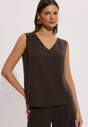 Kobieta ubrana w ciemnobrązowy bezrękawnik z dekoltem w serek i dopasowane spodnie, nosząca duże złote kolczyki koła oraz złoty pierścionek na jasnym tle.