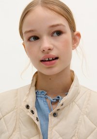 Veste en jacquard crème clair avec des boutons-pression, associée à un blouse bleue avec un col froncé. Fond clair.