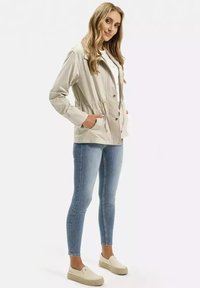 Beżowa lekka kurtka z troczkiem w talii, guzikiem z przodu i bocznymi kieszeniami, noszona z jasnoniebieskimi rurkami i beżowymi butami.