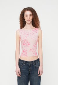 Νέα γυναίκα με σγουρά κόκκινα μαλλιά, φορώντας ροζ εμπριμέ τοπ χωρίς μανίκια και σκούρο μπλε τζιν, στέκεται μπροστά σε απλό λευκό φόντο.