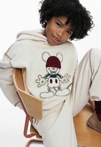 Sudadera crema con costuras rojas, con un diseño de ratón de dibujos animados bordado que lleva un gorro burdeos; combinada con pantalones crema a juego.