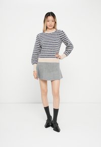 Pullover mit navy und cremefarbenem Muster; hellbeige gerippter Saum. Grauer Rock mit subtiler Textur. Schwarze Schuhe und dunkle Socken runden das Outfit ab.