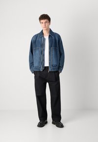 Jachetă din denim într-o nuanță de albastru mediu, având două buzunare mari în față, închidere cu fermoar și o croială lejeră. Purtați peste un tricou alb și pantaloni negri.