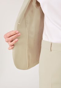 Beiger Blazer aus glattem Stoff mit dezentem Logo-Muster auf dem Futter. Eine Hand, die den Blazer hält, zeigt eine maßgeschneiderte Passform.